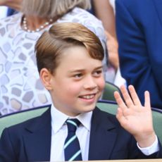 Prince George wears a navy suit and striped tie and waves while watching Wimbledon next to dad Prince William, who is wearing a gray suit