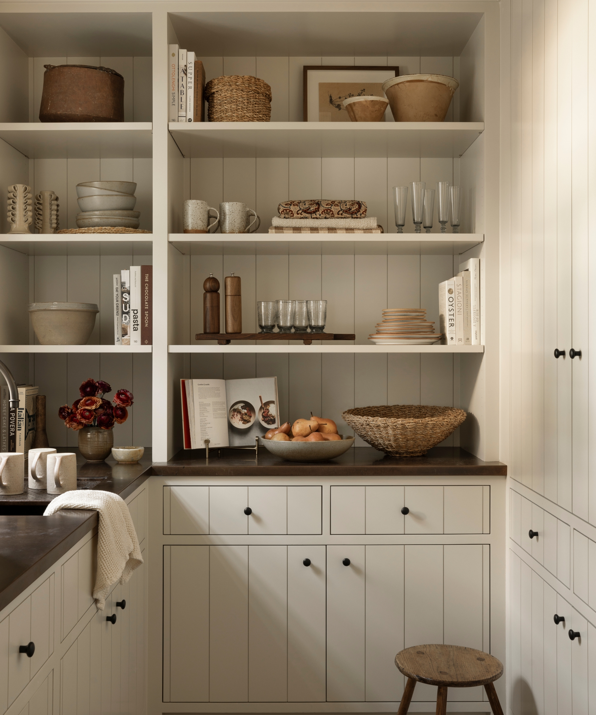 A small walk-in pantry with warm white shiplap style cabinets, dark metal countertops, and open shelving