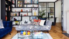 white living room with modular dark wood shelving different levels, books, artwork, vases, chaise, coffee table, blue couch, couch pillows, hardwood floor
