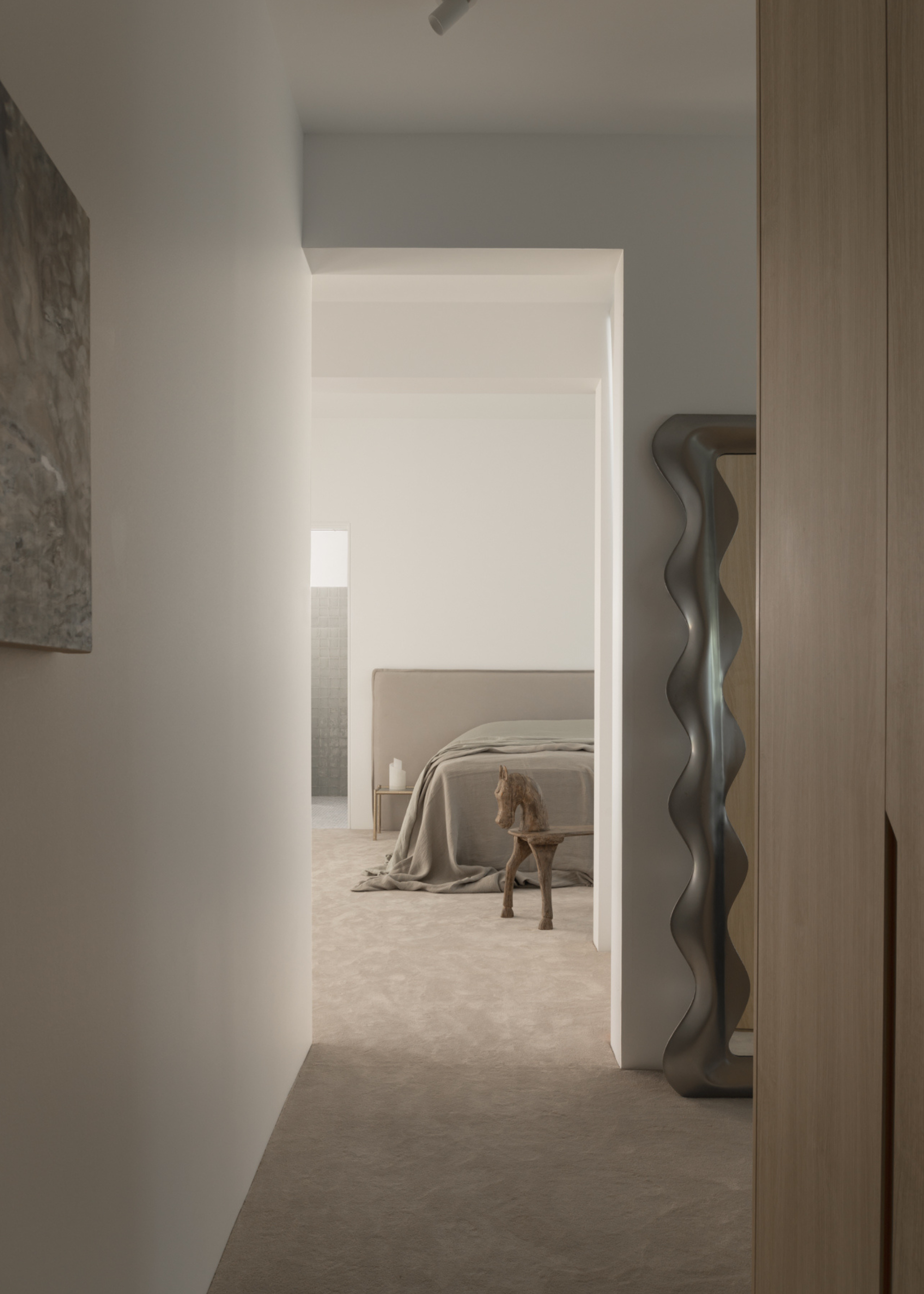 A passageway leading to a white bedroom with a gray bedscape, a wavy chrome mirror, and a horse-shaped wood stool