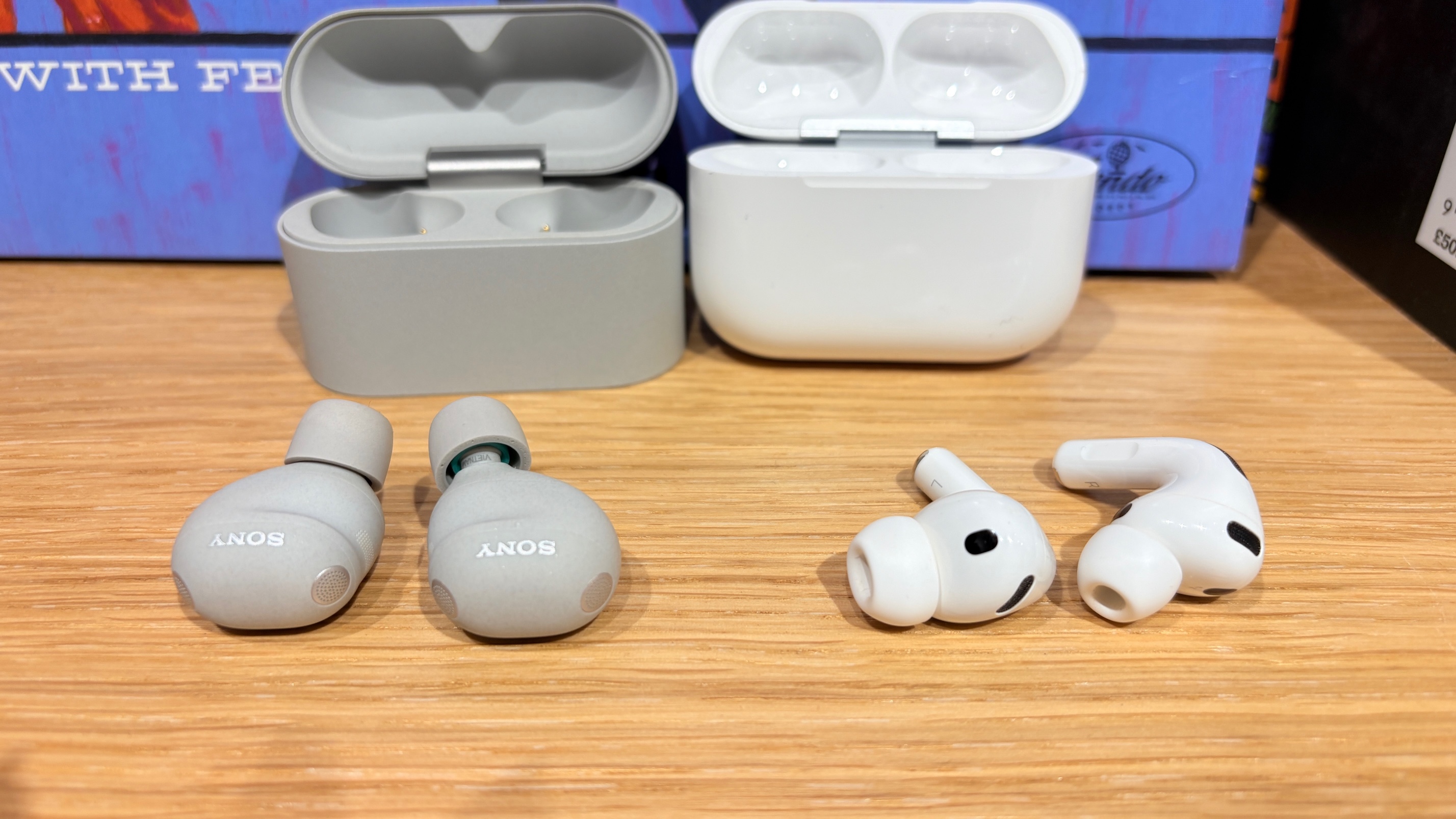 A pair each of the Sony WF-1000XM6 and Apple AirPods Pro 3 earbuds lying on a wooden surface in front of their open charging cases.