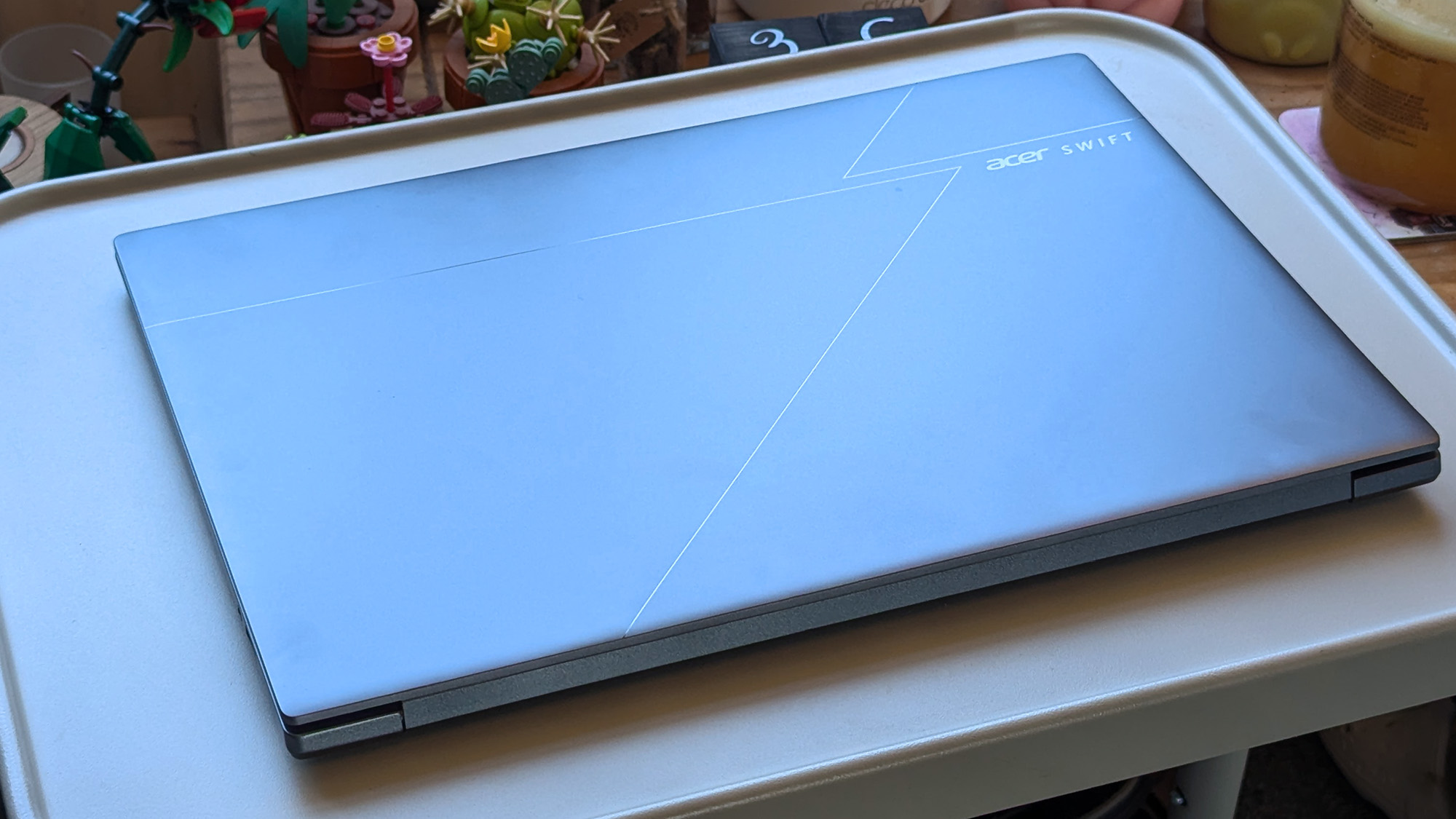 A closed Acer Swift laptop rests on a light-colored tray, its metallic lid featuring angular line accents and the “Acer SWIFT” logo. Small potted plants and colorful décor sit in the background.