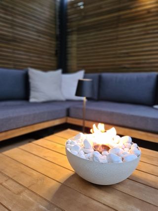 A backyard with a firepit bowl on a wooden table and a grey sofa in the background