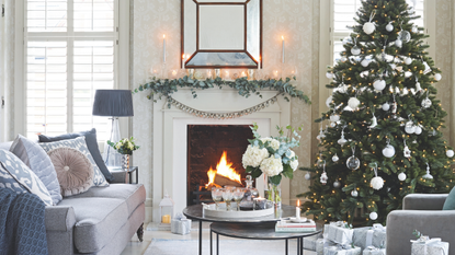 Living room with lit fire and decorated Christmas tree, silver wrapped Christmas presents and decorated mantelpiece with lit candles.