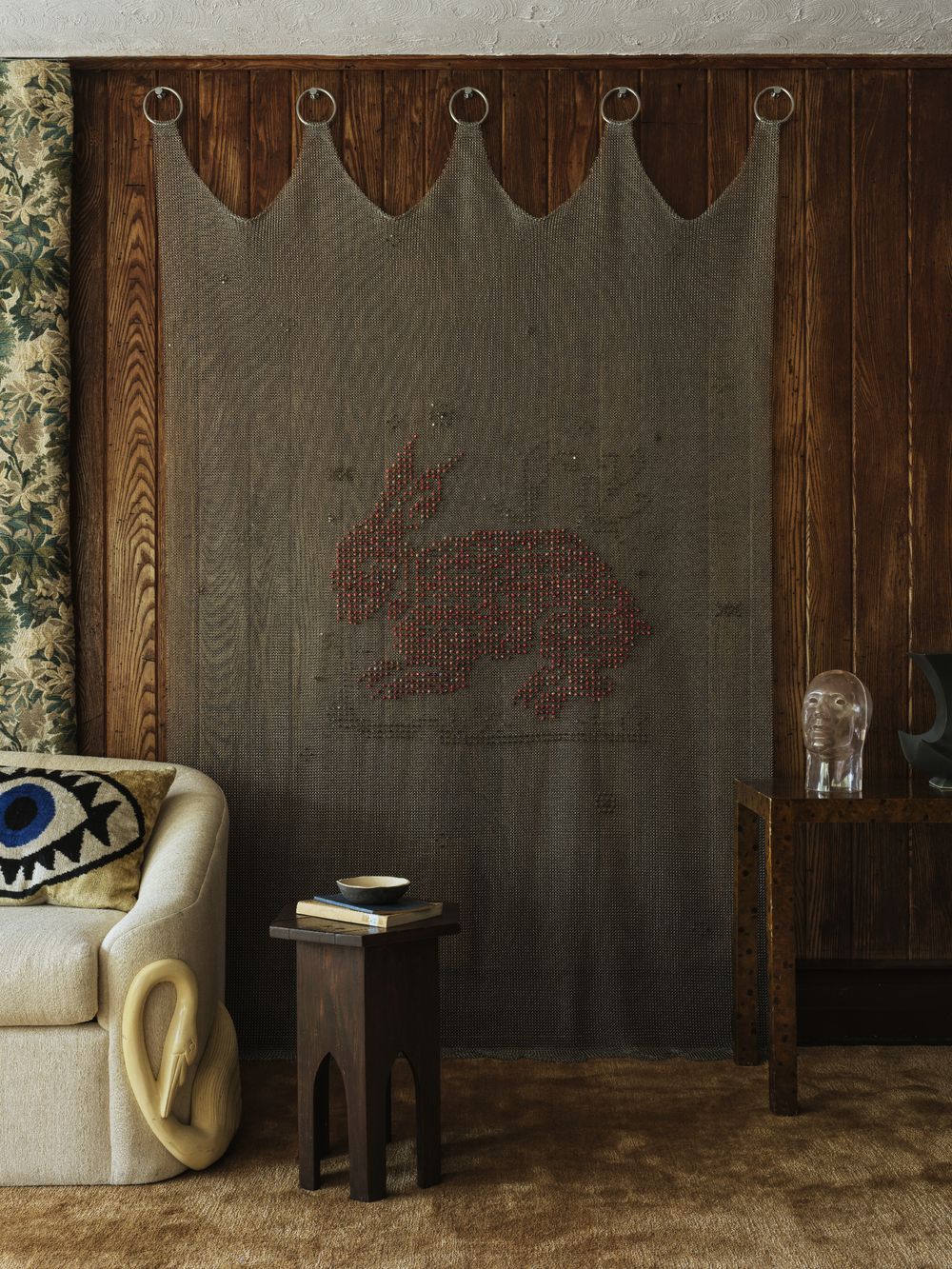 modern living room with silk carpet, corner of beige sofa with pillow with an eye, timber side table with books, timber panelled walls, floral wallpaper, and chainmail tapestry