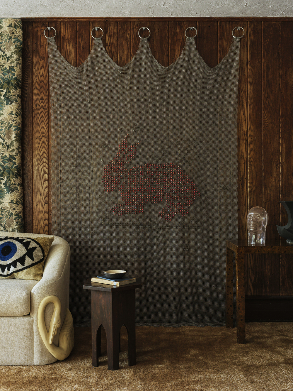 modern living room with silk carpet, corner of beige sofa with pillow with an eye, timber side table with books, timber panelled walls, floral wallpaper, and chainmail tapestry