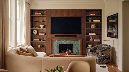 Living room with warm white walls, dark wood media wall joinery, curved beige sofa and Dedar patterned grey and white armchair