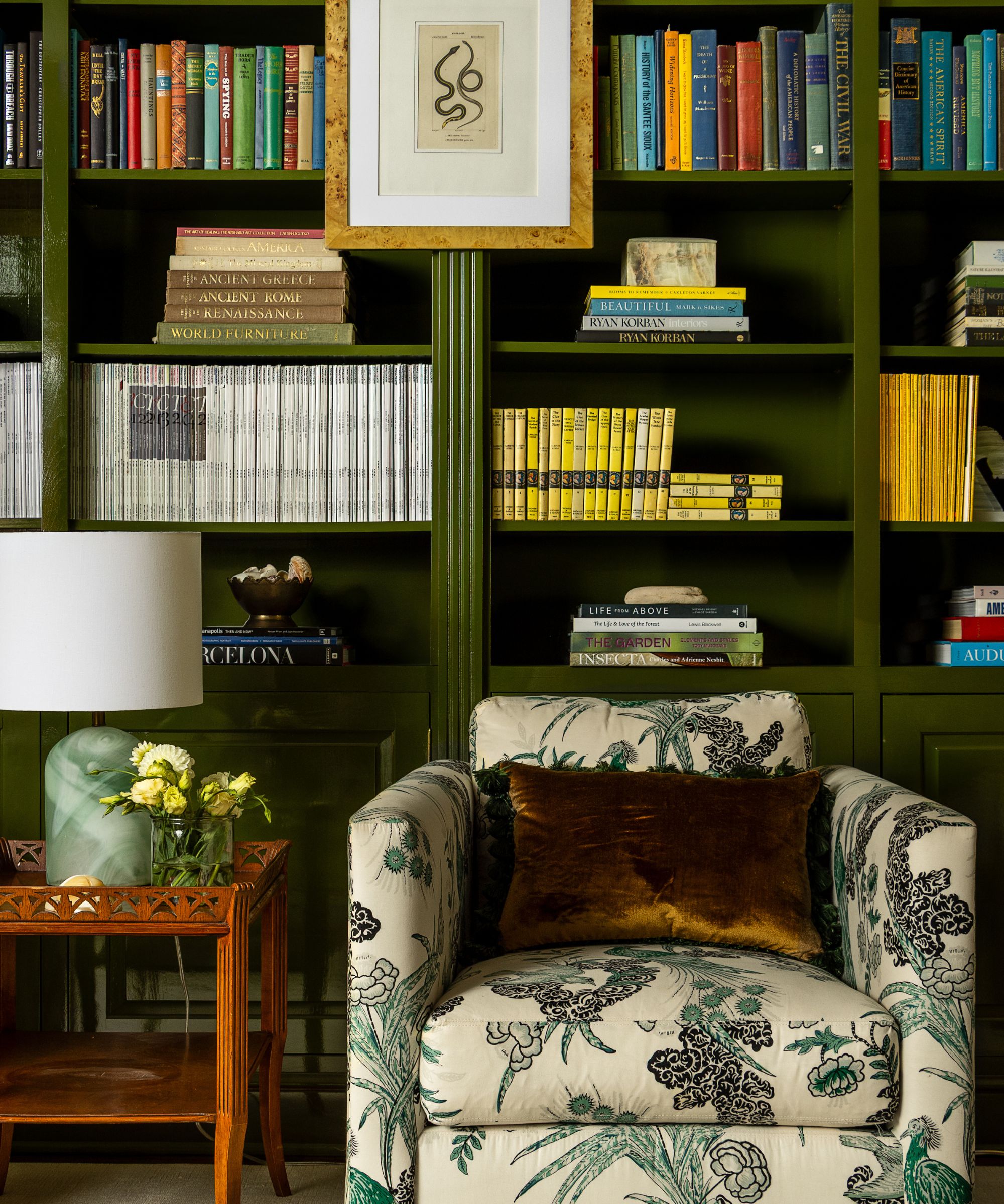 Library with moss green painted bookshelves, floral armchair with dark brown velvet cushion, wood side table with lamp and vase