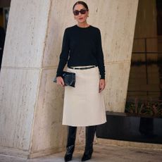 Woman wears black top, white skirt, black belt, black boots, and black clutch while posing for the camera. 