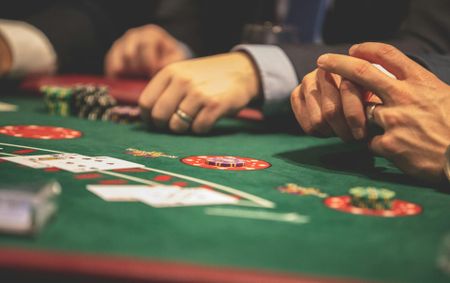 A high-angle, close-up shot of several men's hands at a poker table. The hands are resting on the green felt, with a pile of poker chips in front of them and a few playing cards face up on the table