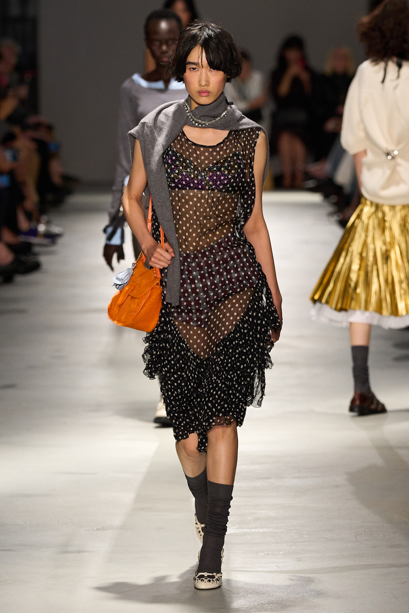 Layering staples for spring 2026 is shown in a photo from No. 21's runway show, with a model walking while wearing a gray cardigan tied around the shoulders with a crystal necklace layered on top that's worn with a sheer polka dot dress with a bralette and briefs, gray knee-high socks, white pumps, and an orange shoulder bag
