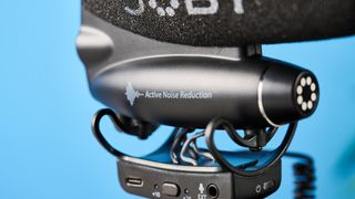 a black shotgun microphone by joby called the wavo pro is photographed by itself against a blue background, showing its muff, dead cat, attachment foot, and connection ports