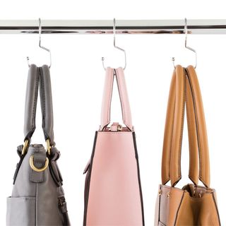 Metal purse hangers hanging from a metal rod with three handbags in different styles in grey, pink and orange hanging from them