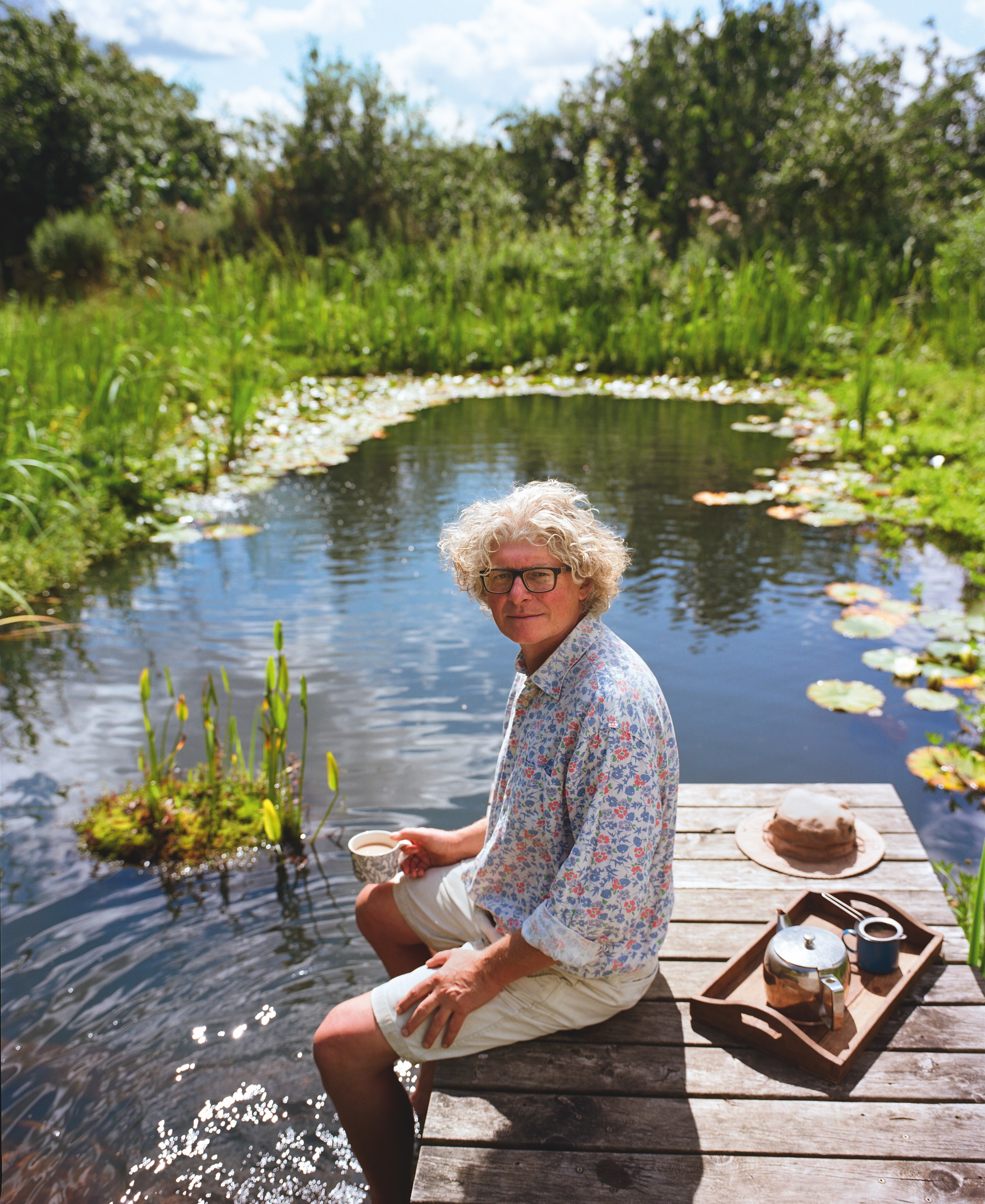 A man in a floral shirt and cream chino shorts, sitting on a boardwalk with his legs dangling in a natural pool