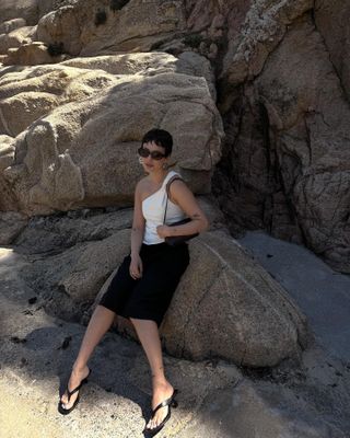 Woman sitting on rocks at beach with a white top, black skirt, and sandals