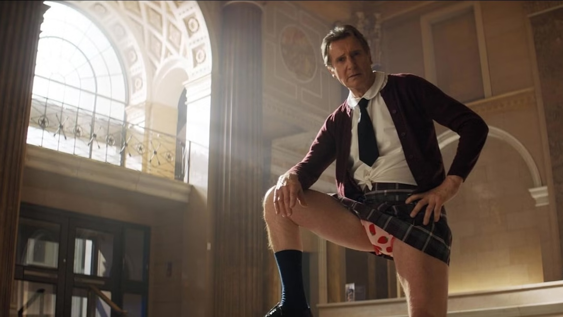 Liam Neeson dressed as a school girl standing in a bank with his leg up 