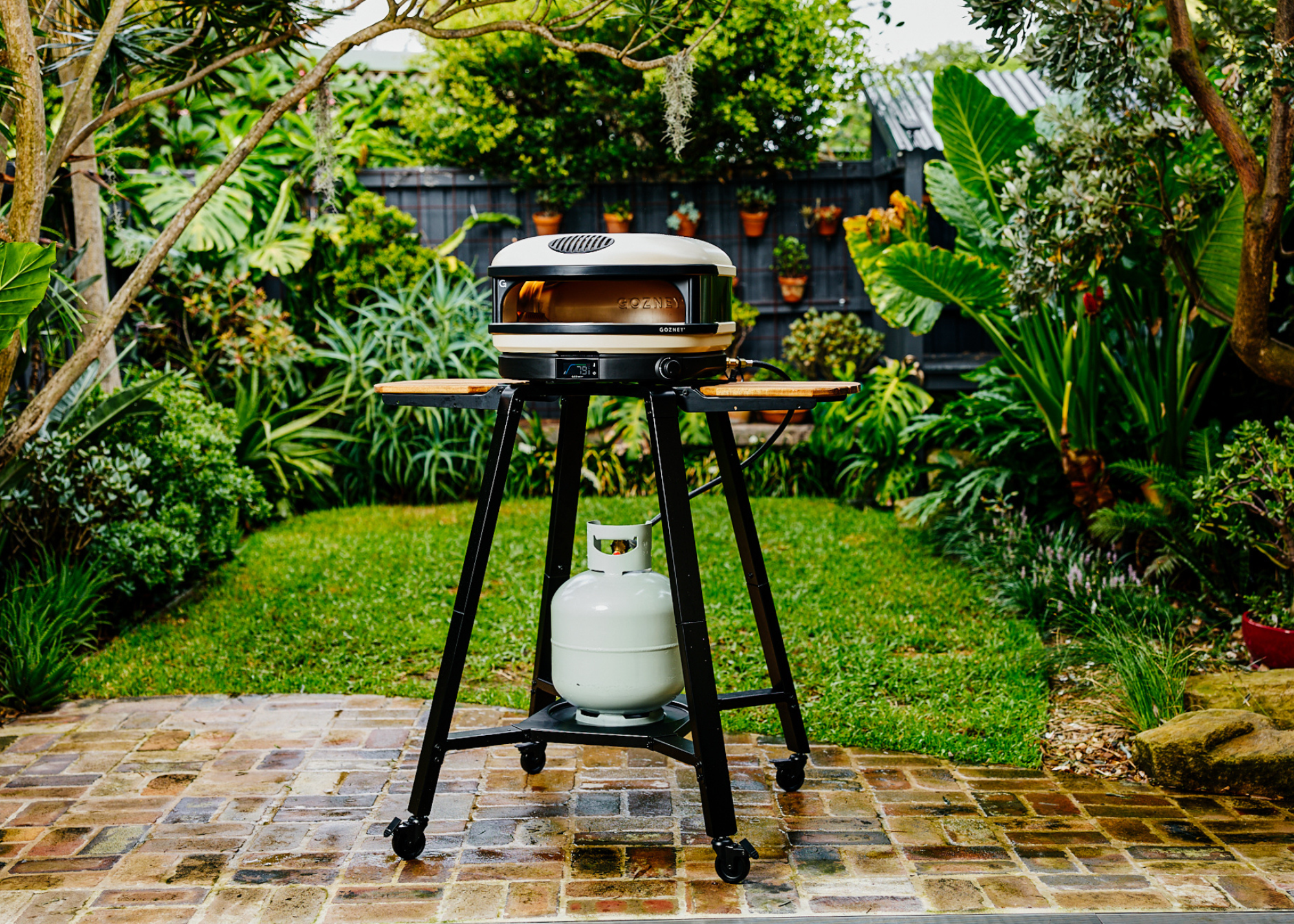 Gozney&#039;s Arc XL pizza oven in a garden patio with grass and plants in the background and pots of plants hung on the black fence behind