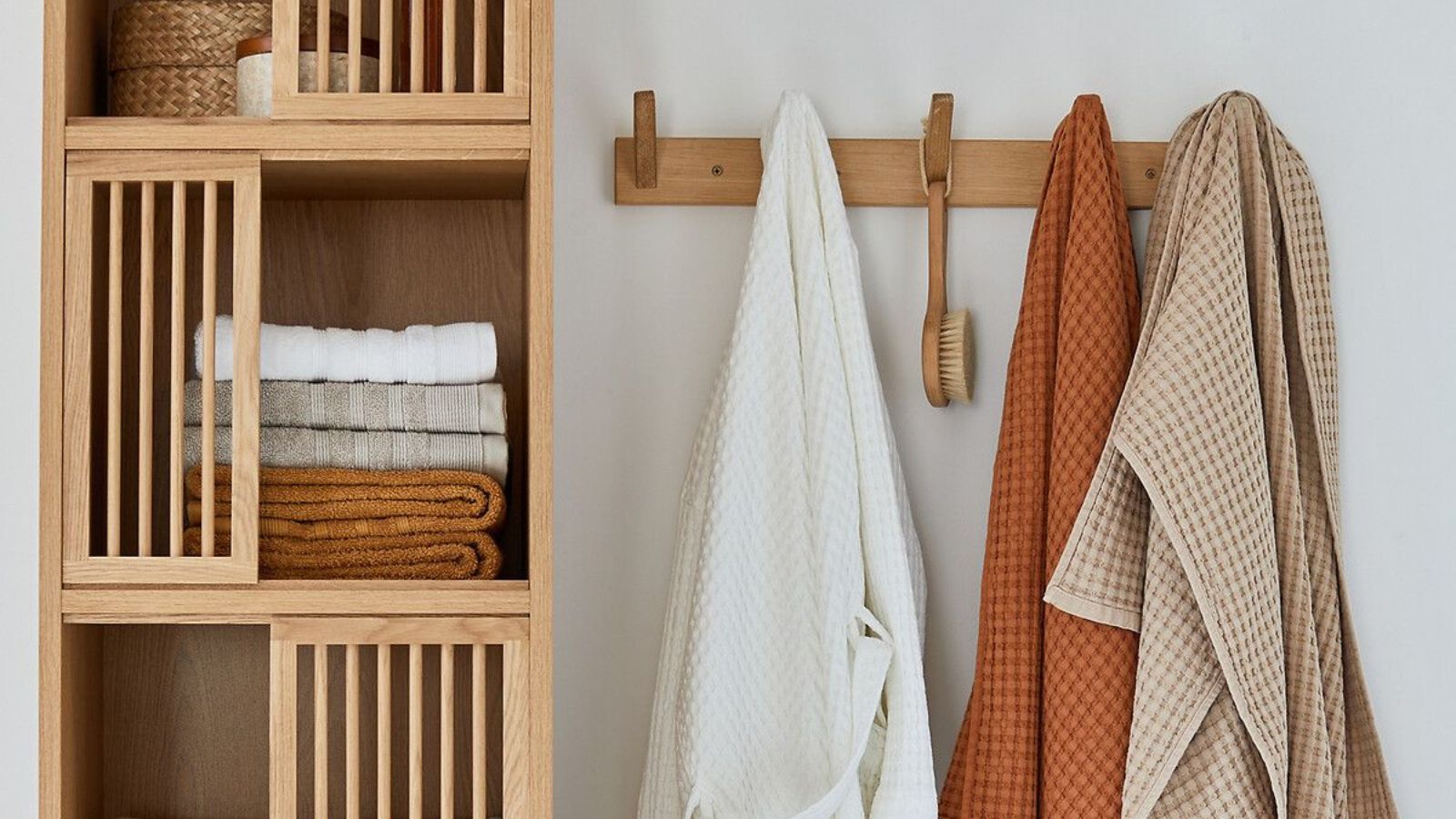 A light oak shelving unit with slatted doors holds neatly folded towels in cream, sage and rust tones. Beside it, a simple wooden peg rail supports a white robe, textured bath towels and a natural bristle body brush.