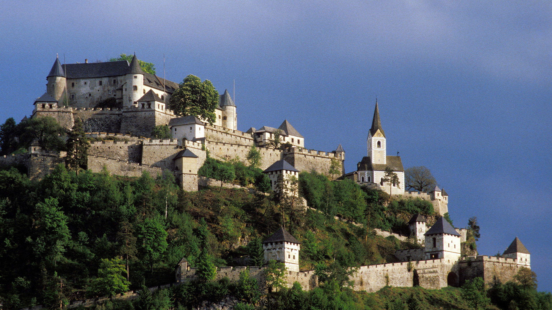A view of a stone castle on the side of a hill, with walls that snake up the side mixed in with trees.