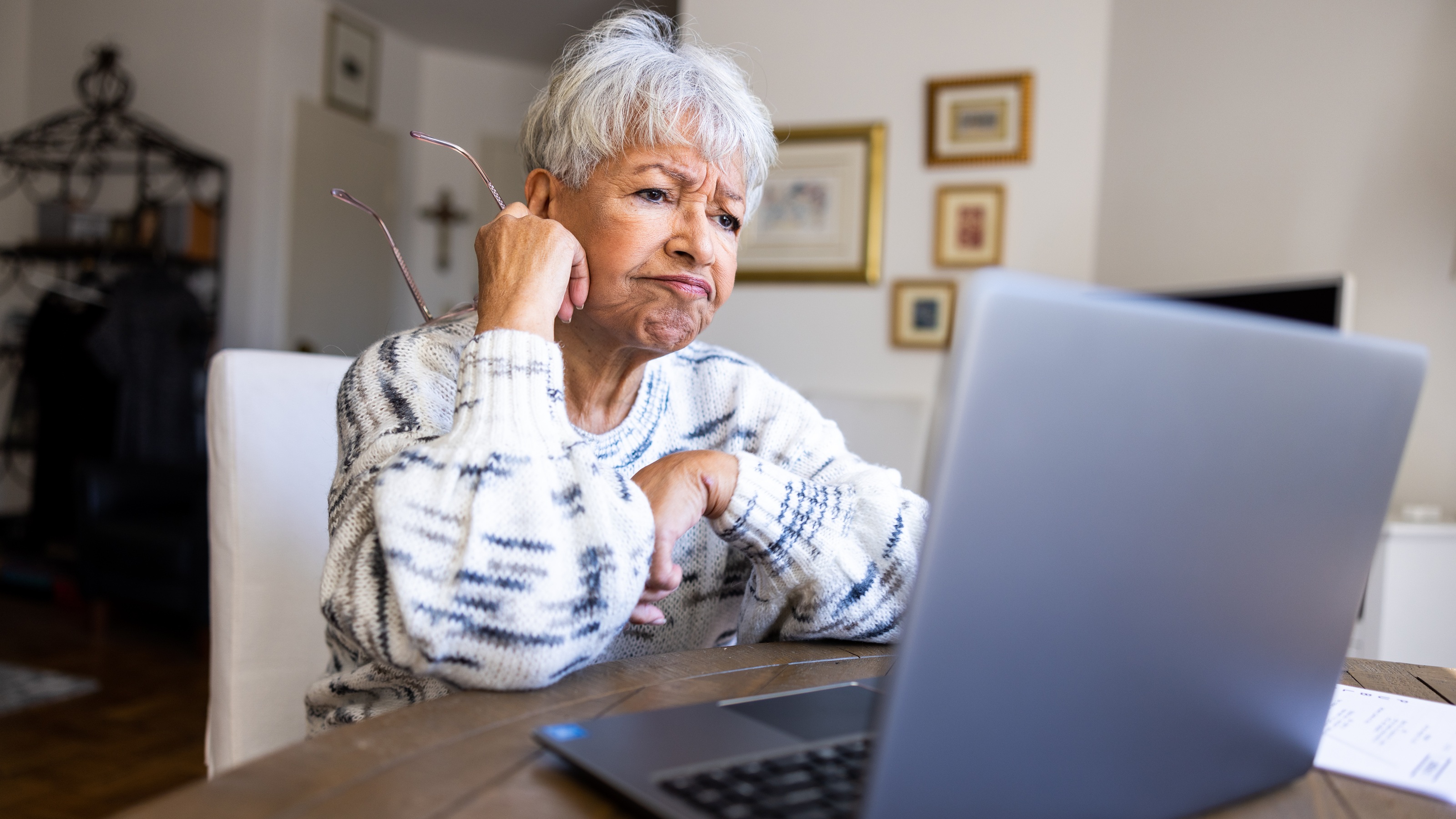 An older woman has an unsure expression on her face as she looks at her laptop at her dining room table. 