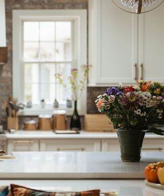 A white kitchen counter with a dark green vase of colorful flowers, with a counter with wooden canisters, a black vase, a gray brick wall with a large white window and white wall cabinet behind it