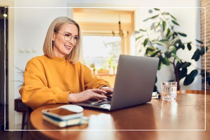 Blonde woman in ochre jumper working on laptop on dining table at home