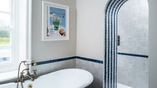 A blue and white tiled bathroom with large bathtub with gold faucets, colorful artwork, and tiled archway to a shower behind