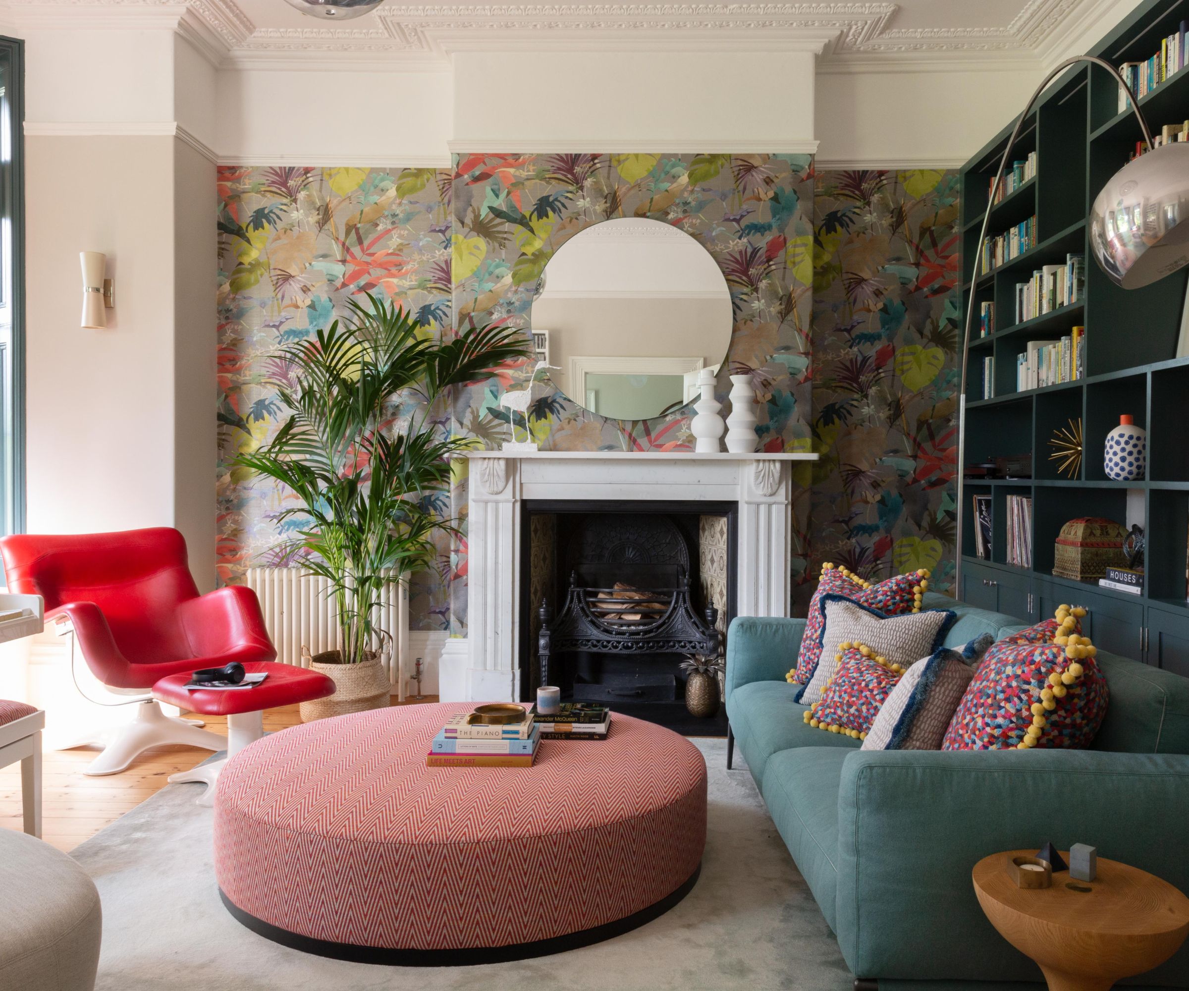 Bright, eclectic living room with a white fireplace and ornate ceiling cornicing. A large round mirror sits above the mantel, backed by colourful botanical wallpaper. A teal sofa, red armchair, patterned cushions, bookshelves and a pink circular ottoman create a lively, welcoming space.
