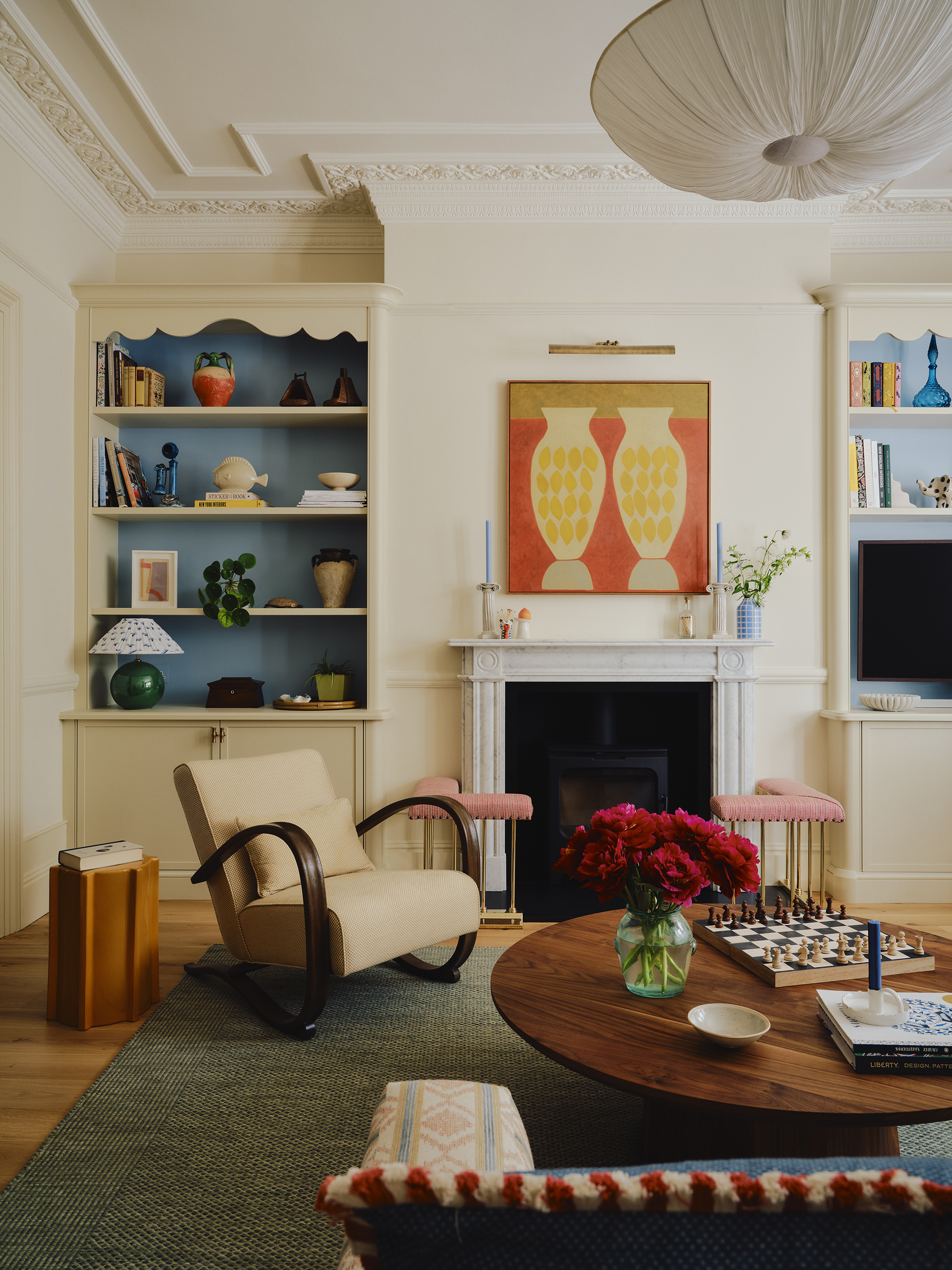 Image of a bright, playful living room design. The walls are white, and there are built-in bookcases painted blue on either side of the fireplace. There is a round wooden coffee table with a chessboard on it and a cream accent chair.