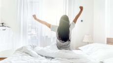 Woman with arms in the air stretching on her bed with back to the camera as she wakes up from sleep