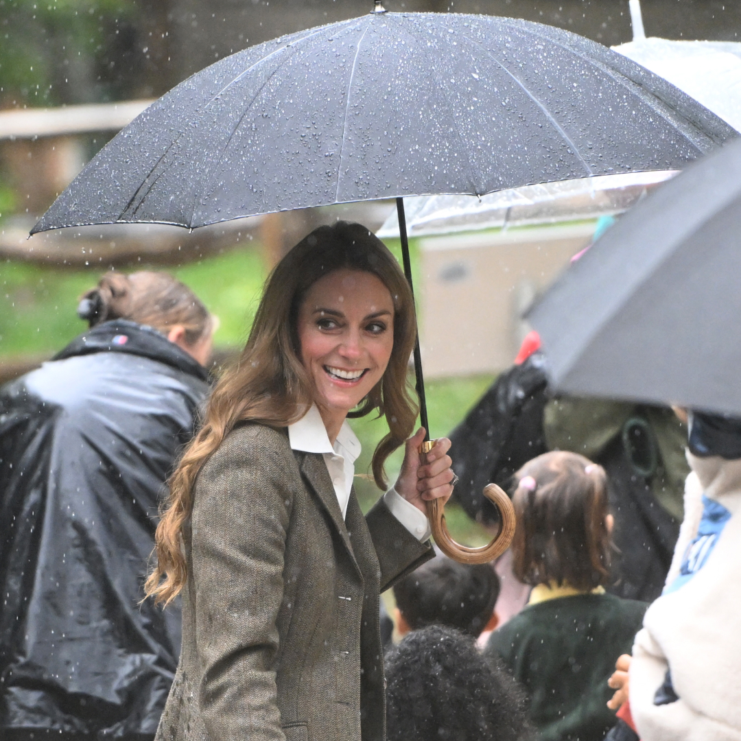 Kate Middleton holding a black umbrella in the rain surrounded by people
