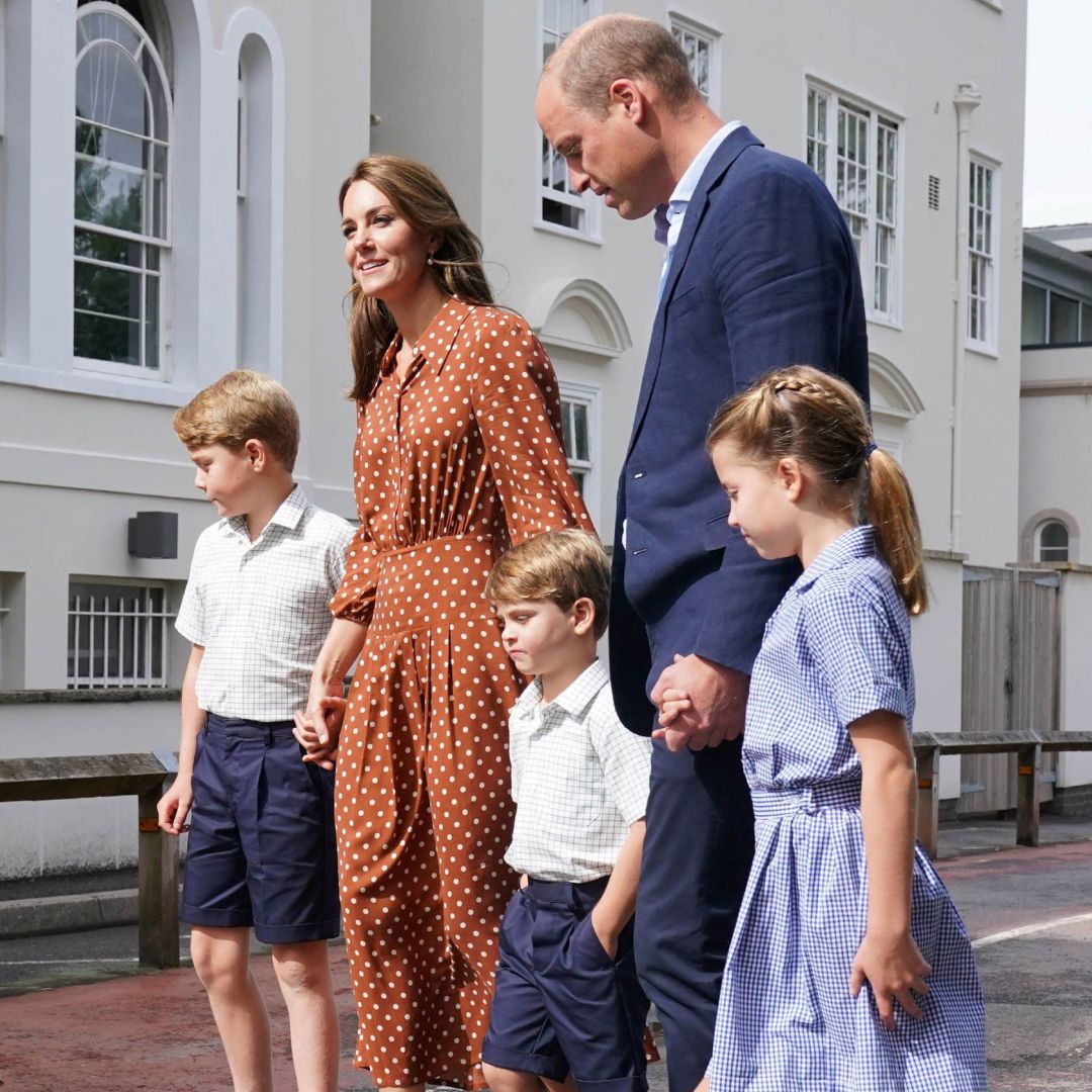 Princess Kate wearing a brown polka dot dress and Prince William wearing a blue suit holding hands with Princess Charlotte, Prince George and Prince Louis, who are wearing navy and white school uniforms.