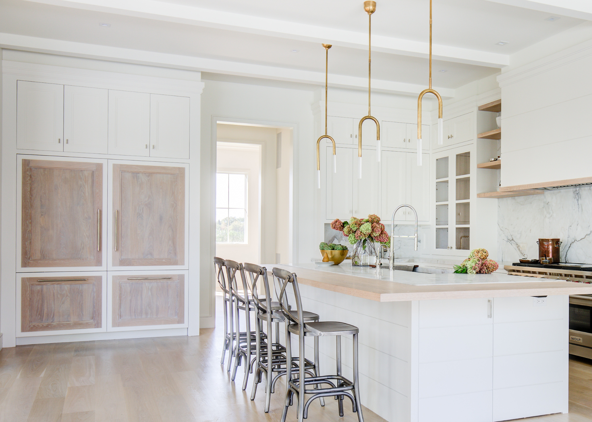 A spacious light white kitchen with a light wood accent, brass island pendants and marble countertops