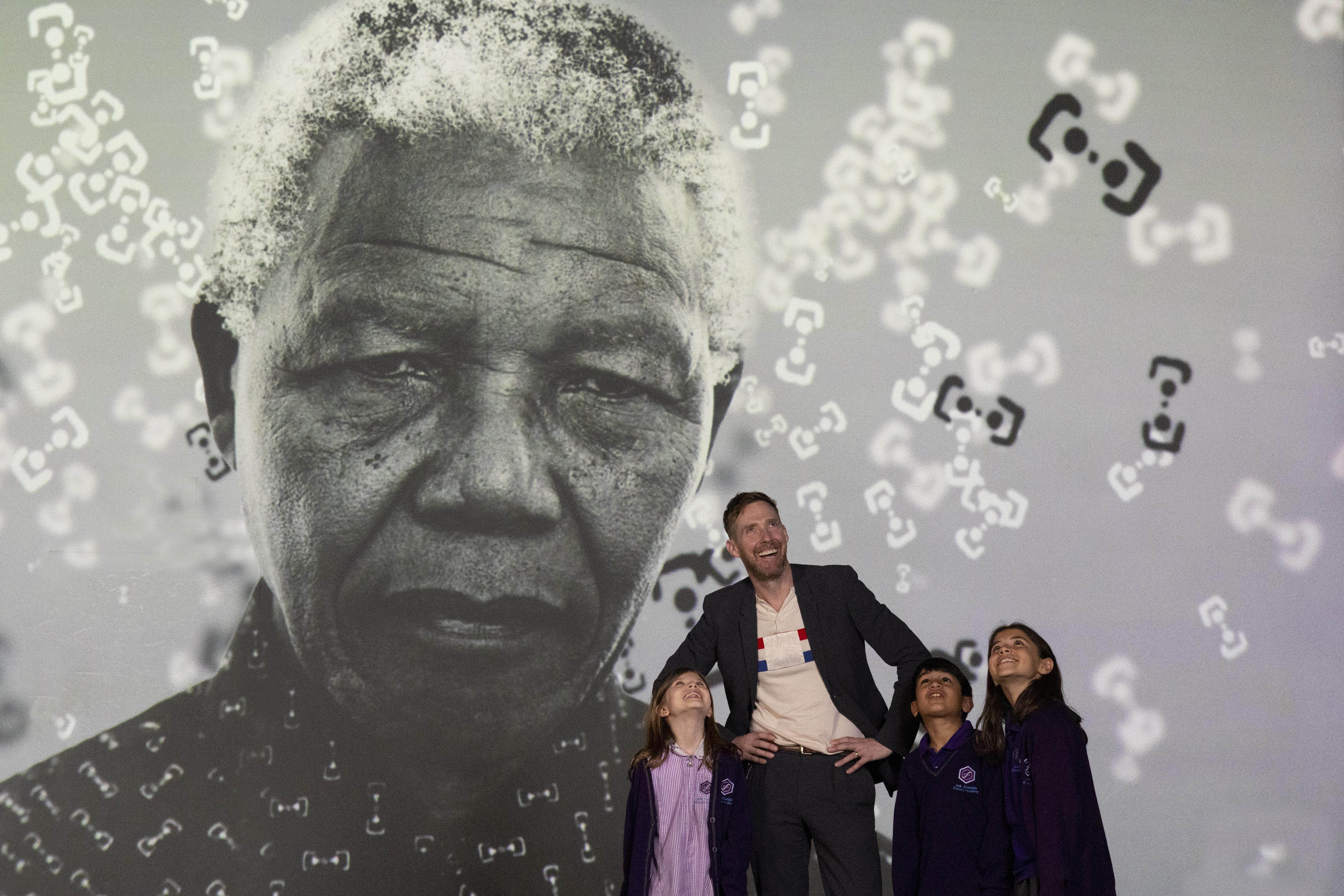 Singer Ricky Wilson crouches alongside three schoolchildren in front of a large-scale projection of Nelson Mandela's portrait at the FRAMELESS London preview, all looking upward in wonder.