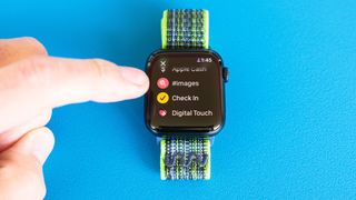 Detail shot of an Apple Watch SE on a bright blue background showing the steps to set up and send a Check In notification in watchOS 11 