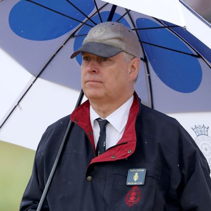 Prince Andrew stands under a white and navy umbrella in the rain while on the golf course, he wears a baseball cap and a navy jacket with a red trim