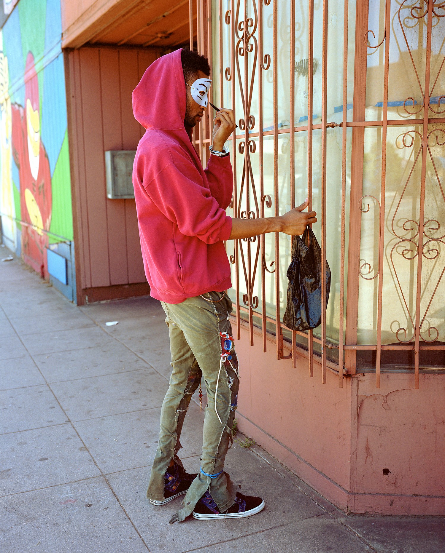 A person in a pink hoodie and ripped pants applies makeup to a white mask while holding a plastic bag next to a barred window on a sidewalk.