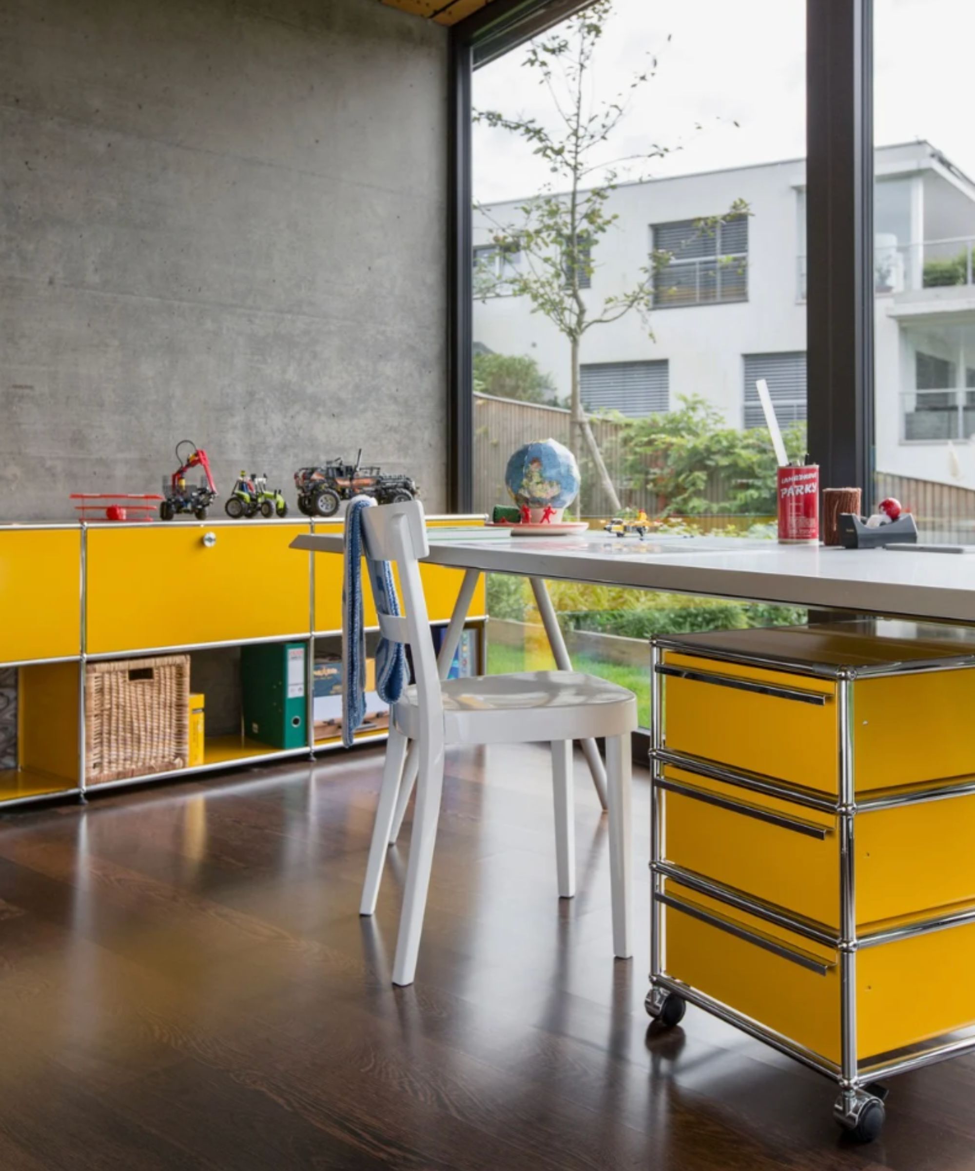 Playful home workspace with bold yellow modular furniture, a white desk and chair, dark wood floors, and large windows overlooking a modern garden.
