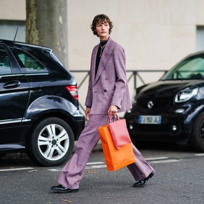 A model wears a black turtleneck pullover, a necklace, a purple checked oversized blazer jacket, a red leather bag, flared suit pants, black leather pointy shoes, outside Hermes, during Paris Fashion Week - Womenswear Spring Summer 2021 on October 03, 2020 in Paris, France.