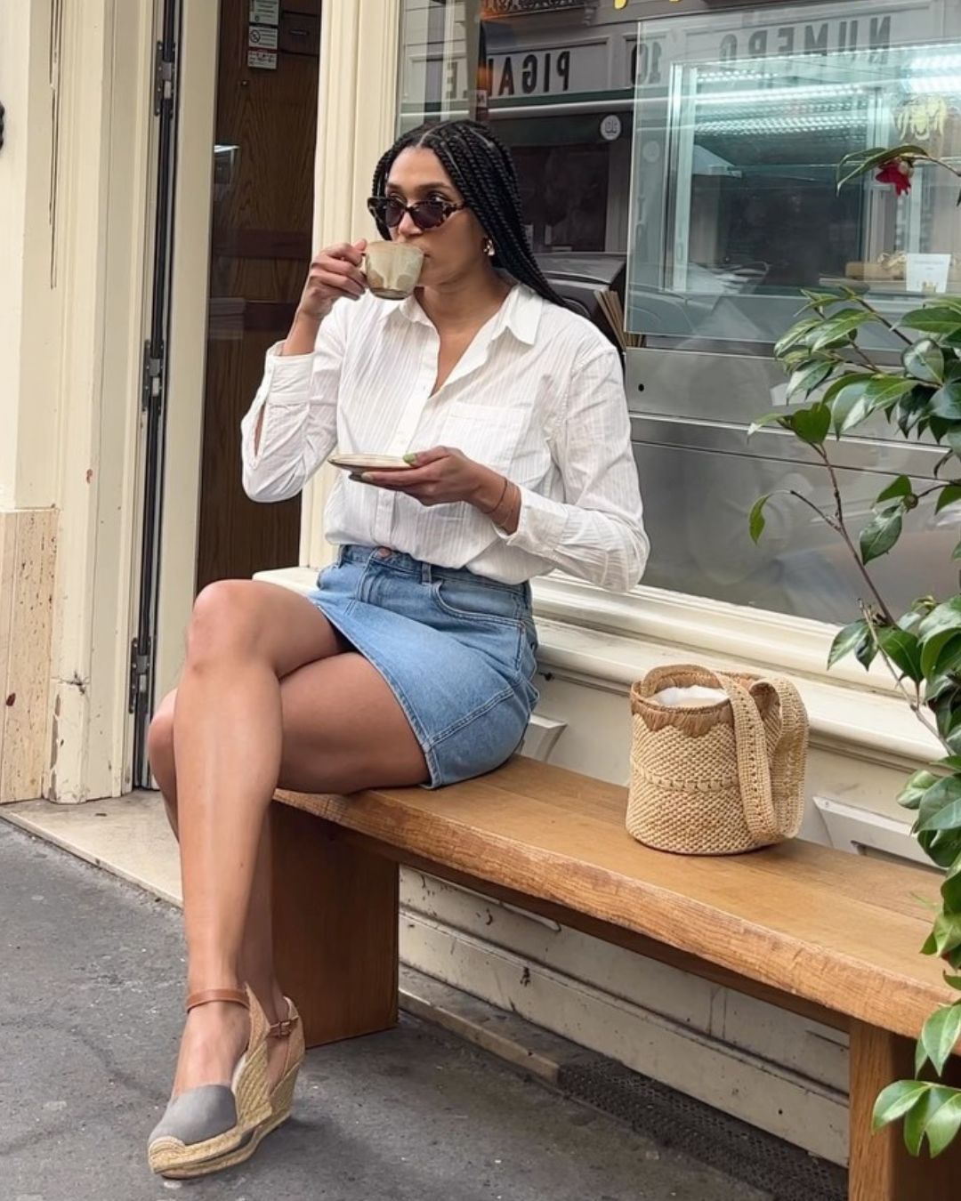 Influencer @lenafarl sits on a bench outside of a coffee shop wearing a white shirt, a denim skirt, and espadrille wedges.