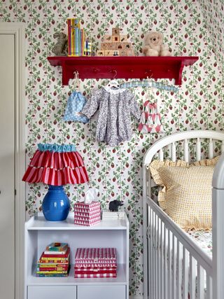 baby girl nursery with cherry wallpaper, red shelf, side table with bright fabric lamp/shade, white cot, clothes hanging up