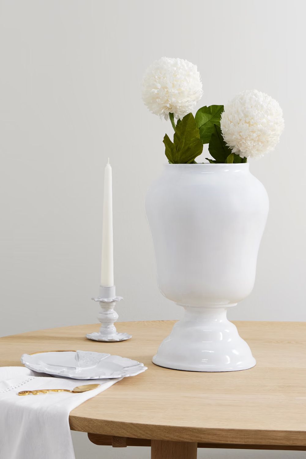 White ceramic footed vase with white flowers in it and a white taper candle beside