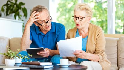 A retired couple sit on a couch looking at paperwork.