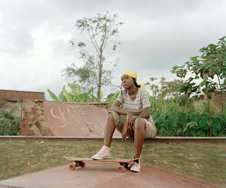 An African woman sitting on a skateboard ramp with one foot on a skateboard.