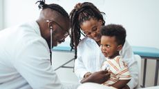A happy toddler boy sits in his mother' lap as a pediatrician leans over with a stethascope to listen to the child's heart