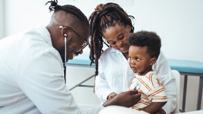 A happy toddler boy sits in his mother' lap as a pediatrician leans over with a stethascope to listen to the child's heart