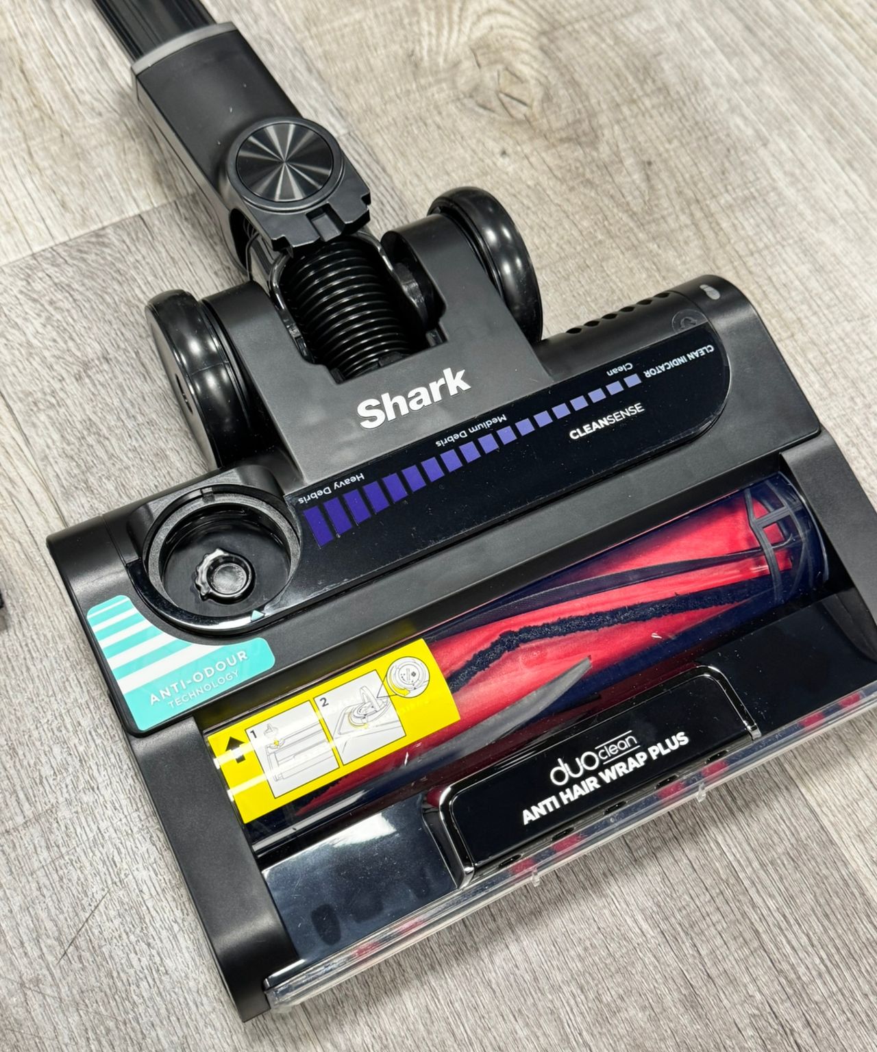 Closeup of the Shark Stratos Cordless vacuum's DuoClean floorhead on laminate floor, showing red bristled roller and Clean Sense IQ indicator.