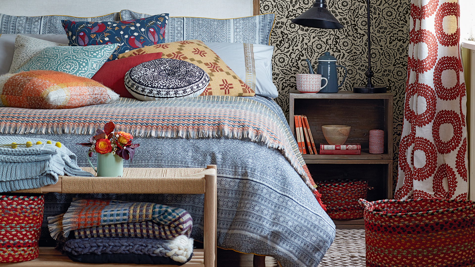 Cosy bedroom with a bed piled high with cushions and throws with patterned wallpaper behind the bed