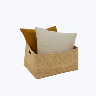 A long hand woven seagrass storage basket with two pillows inside against a white background