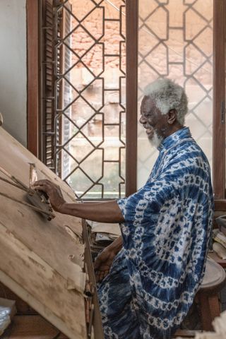Photograph of Demas Nwoko working in his studio, dressed in a long blue and white pattern over shirt, wood framed and pattern window, grey wall, wooden working board, wooden stool